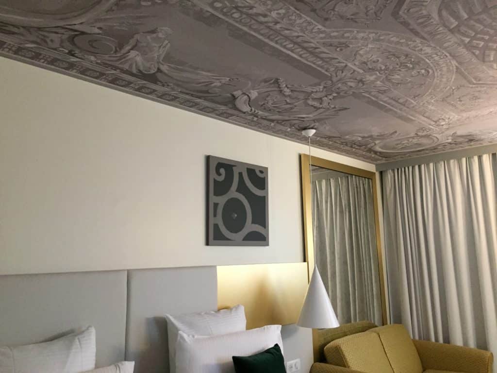 Photo du plafond d'une des chambres de l'Hôtel Le Louis avec un visuel représentant une peinture d'un plafond avec moulures